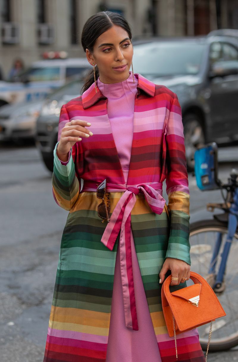 Street-style outfit with a pink dress, multicolored belted coat, and orange bag